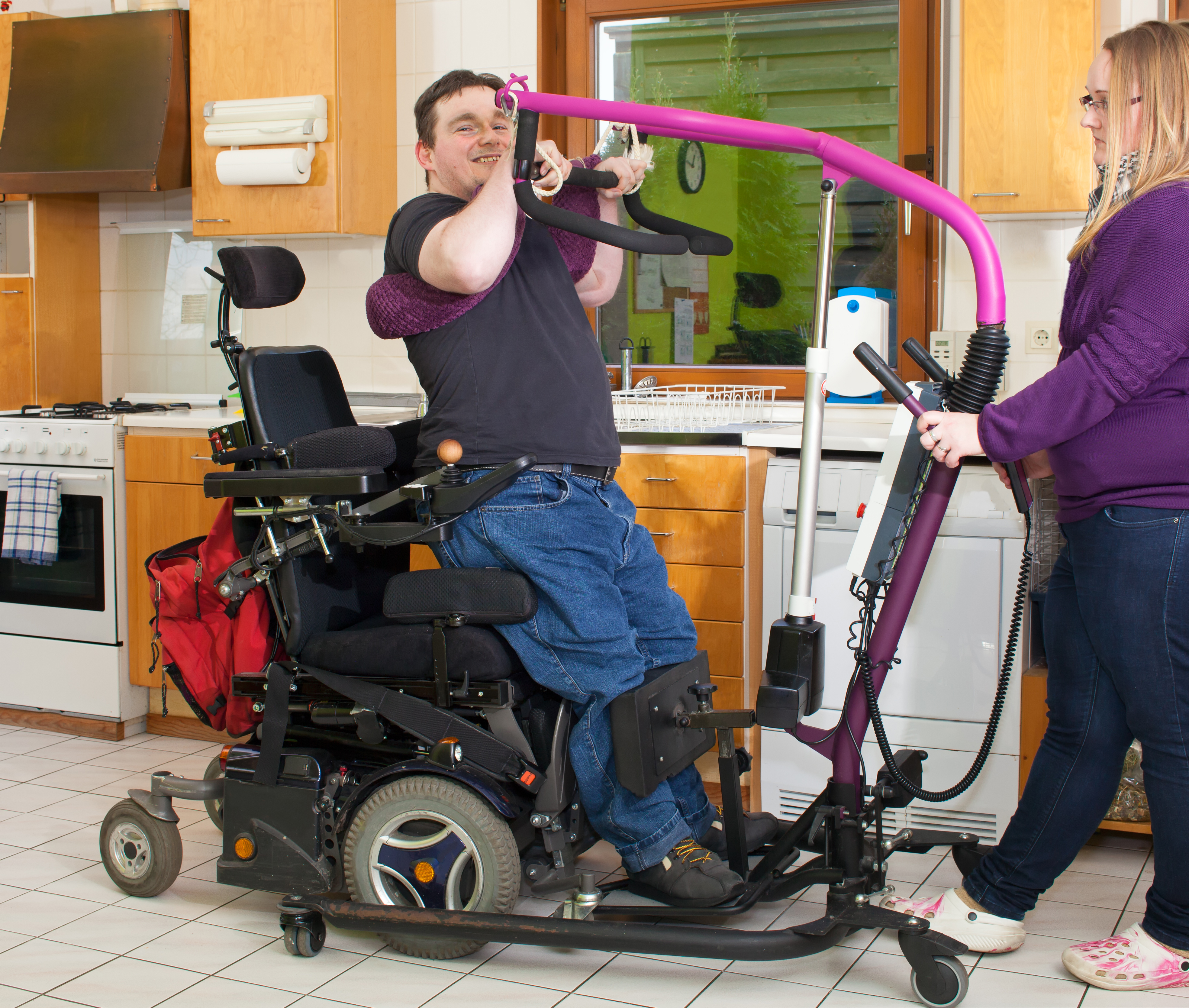 A man in a wheelchair is standing up using lifting equipment, with a provider standing nearby