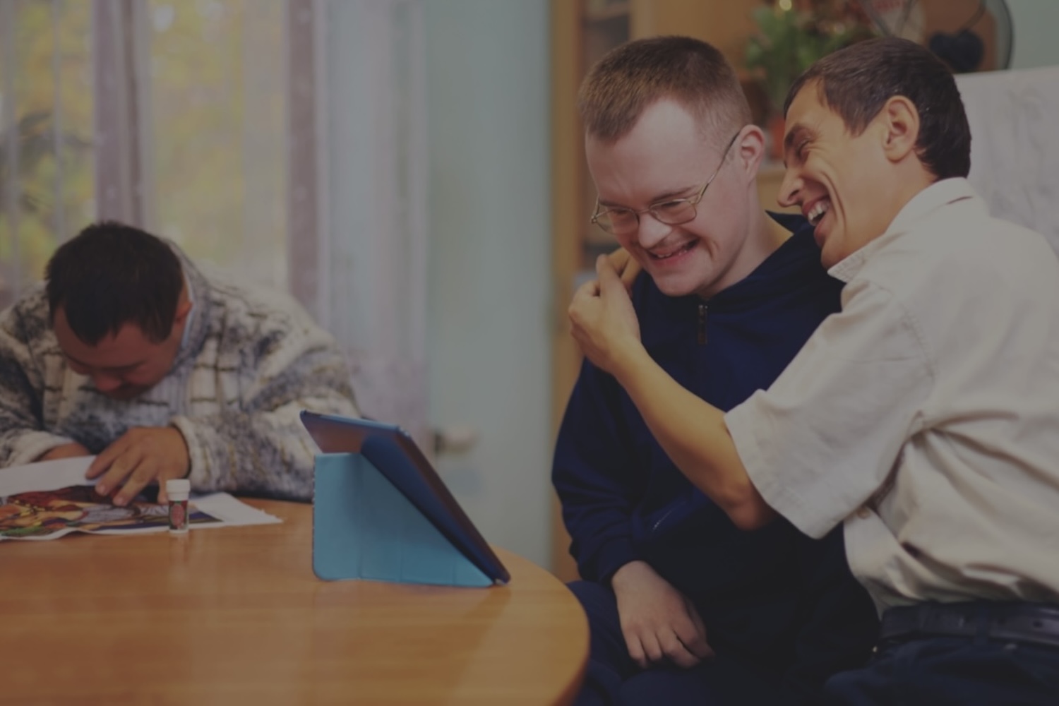 Two disabled men give each other a hug and laugh while viewing something on an iPad screen.