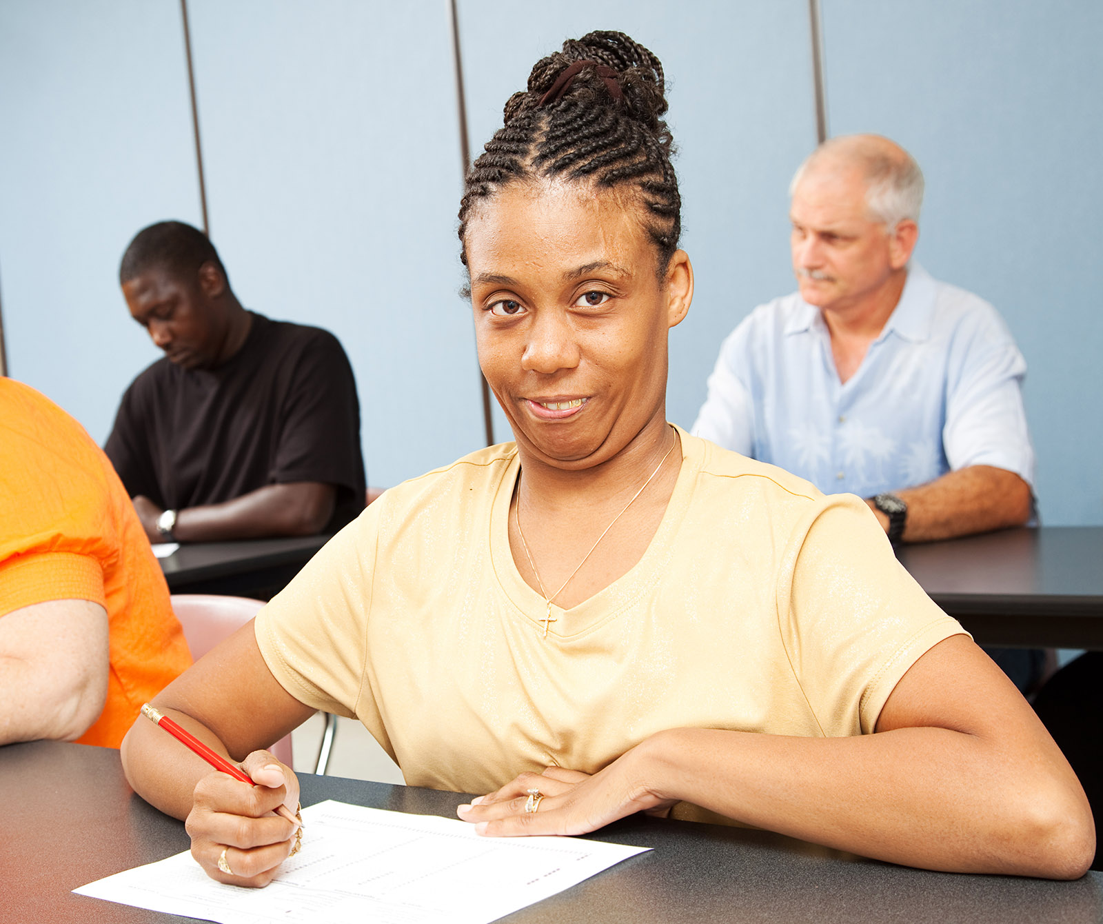 An African-American woman in a yellow t-shirt sits at a table while taking notes on a piece of paper.